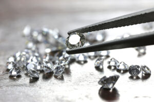 Several loose round cut diamonds on a gray table with one held in tweezers.