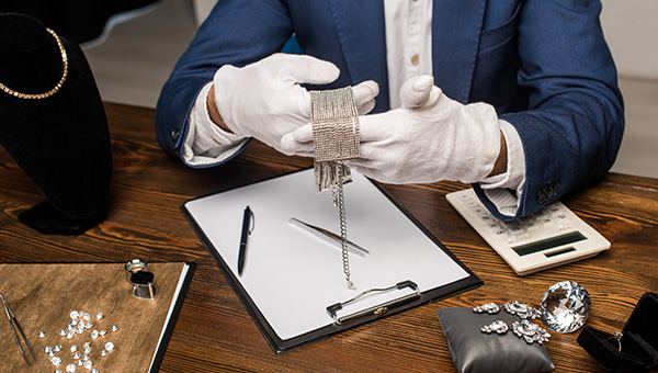 a photo of a gemologist examining a diamond bracelet with his 2 hands wearing white gloves on a wooden table