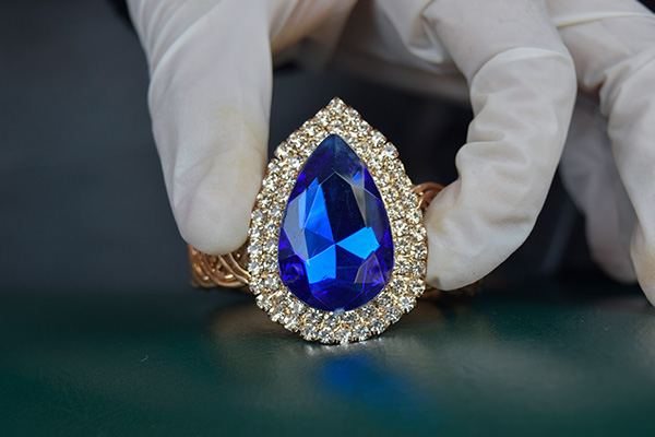 a close up image of a pear cut sapphire with a halo of diamonds around it. the sapphire and diamond piece is being held in white gloves.