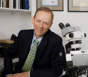 Professional gemologist in dark suit and green tie sitting at desk with binocular microscope in laboratory office