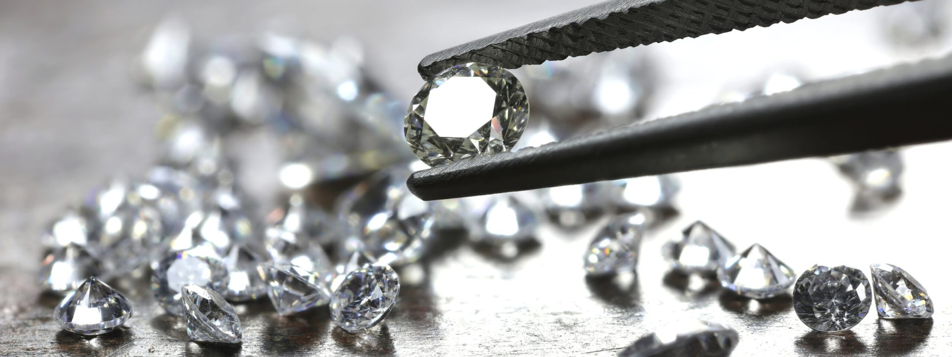 Several loose round cut diamonds on a gray table with one held in tweezers.