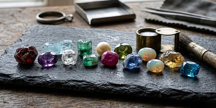 a close-up picture of uncut birthstones sitting on a black slate, a loupe sits besides the stones, hinting that they are being inspected by a gemologist.