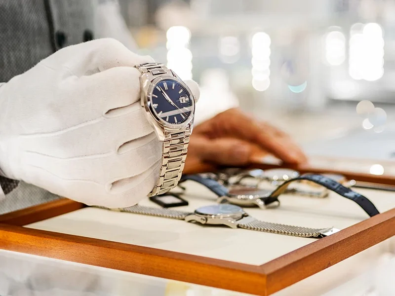 Close-up of a jeweler wearing a white glove holding up a watch for inspection beside a small watch case containing several other watches, resting on top of a glass jewelry display case.